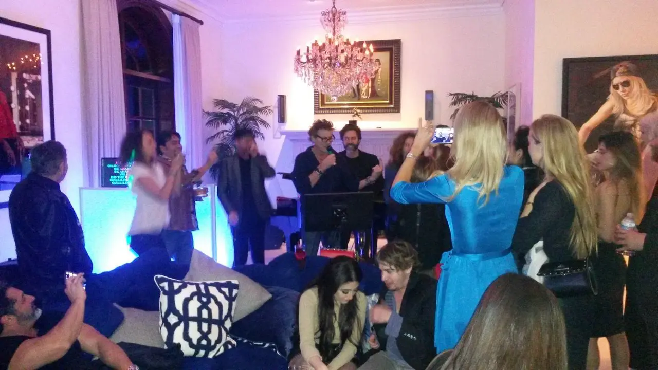 Famous Actor Karaoke A group of people gathers in a living room, some standing and others sitting, with colorful lighting and a chandelier overhead. A woman in blue is taking a photo of the crowd.