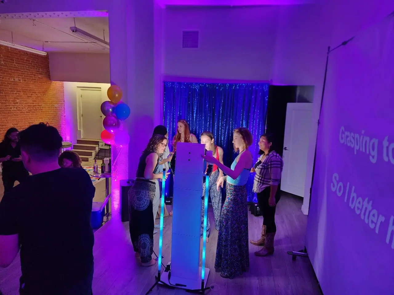 Photo Booth Fun A group of people stand around a lighted photo booth at an indoor party with purple lighting; a karaoke screen is visible on the right.