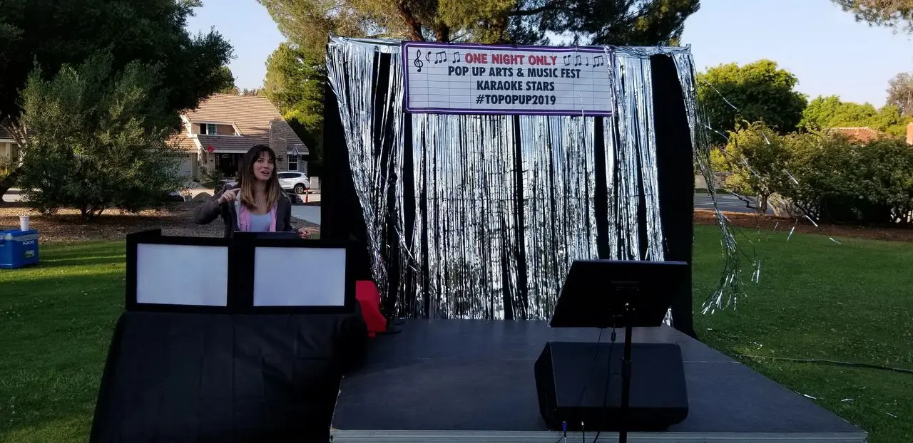 Karaoke dj movie night A woman stands behind a DJ booth on an outdoor stage decorated with silver streamers and a sign for a pop-up arts and music fest called “Karaoke Stars #TOPOPP2019.”.