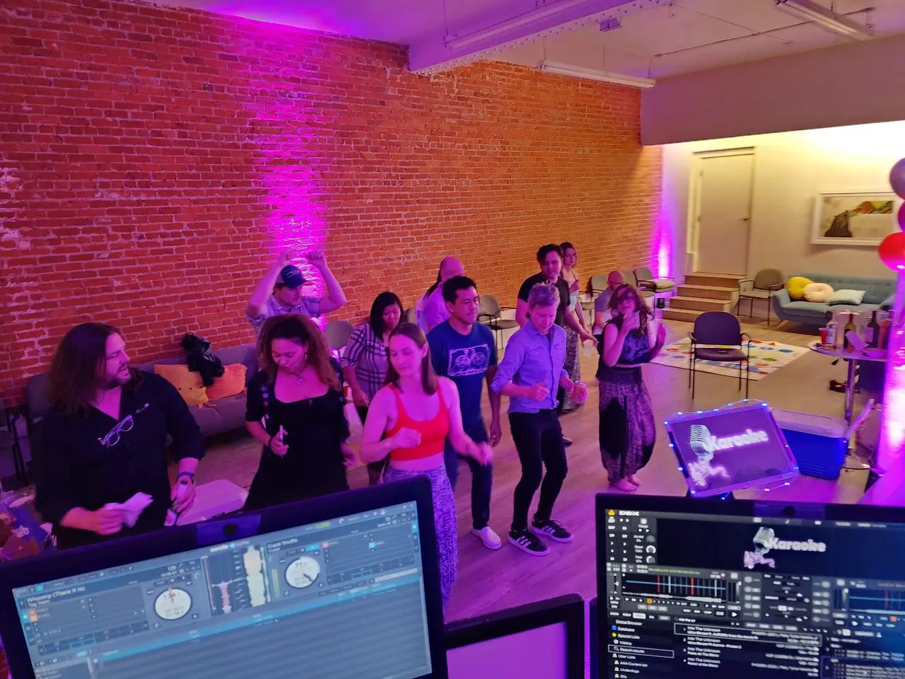 Party Dj Karaoke Party A group of people dance in a room with brick walls and purple lighting, viewed from behind a DJ booth with music equipment and karaoke screens.