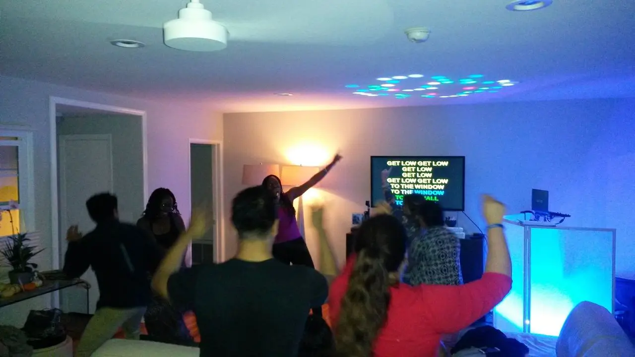 Mr Dj Services Karaoke Dj A group of people dance in a living room with dim lighting while karaoke lyrics are displayed on a TV screen in the background.