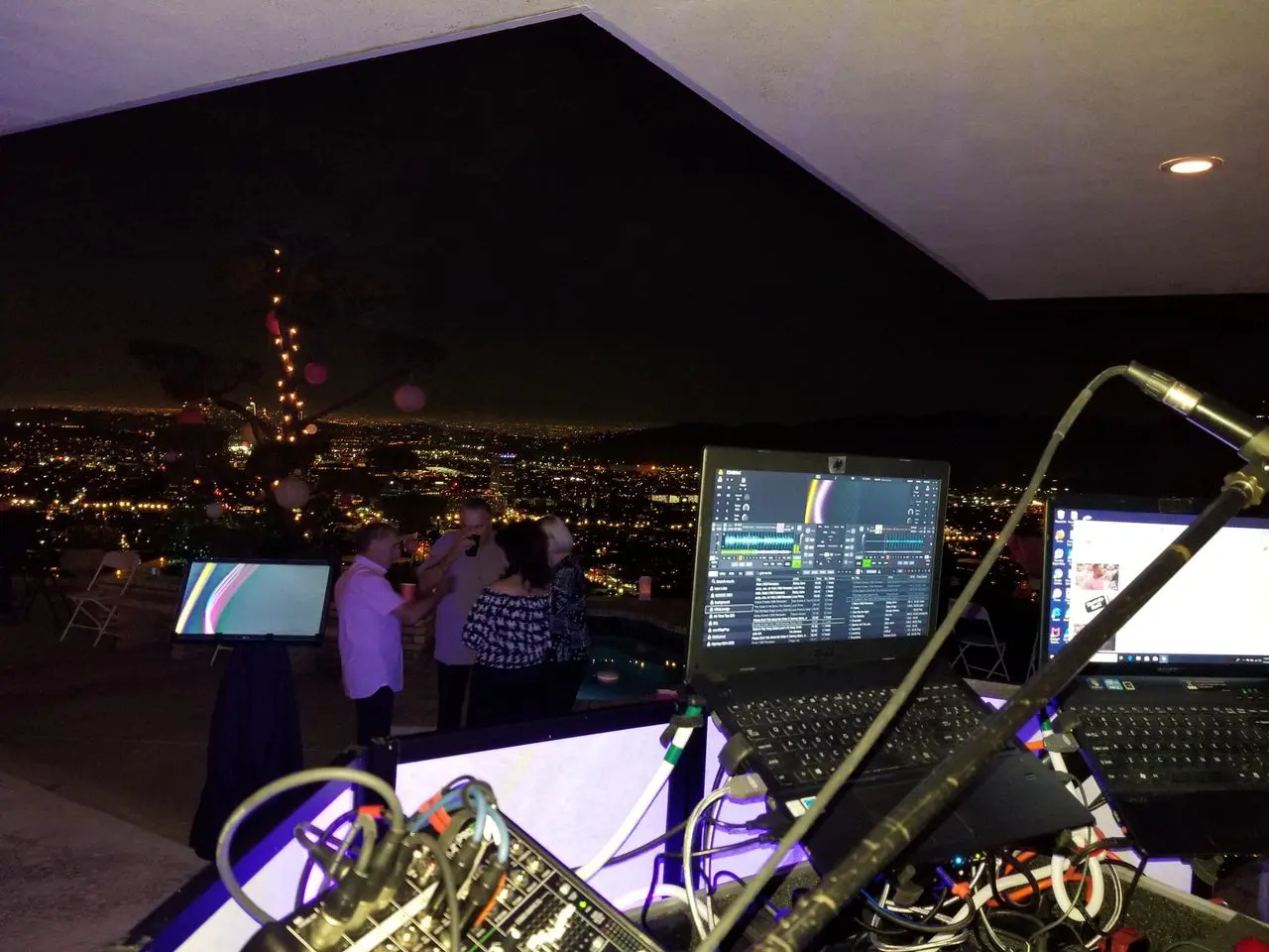 Mr Dj Services View from a DJ booth overlooking a city at night, with a laptop, cables, and microphones in the foreground and people dancing in the background.