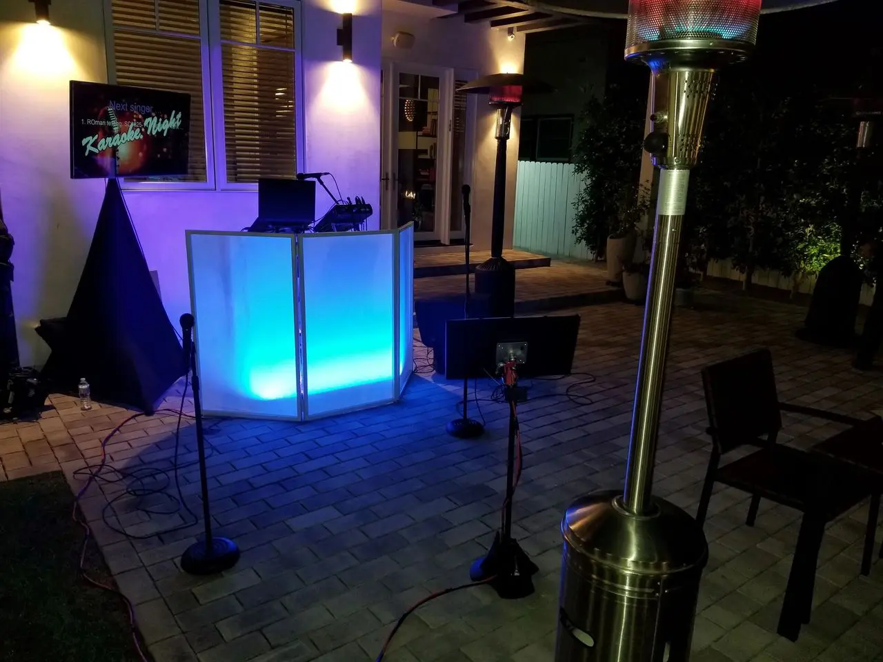 rs=w_1280 (6) Outdoor karaoke setup with a glowing DJ booth, microphones, speakers, and patio heaters on a tiled patio at night. A "Karaoke Night" sign is visible in the background.