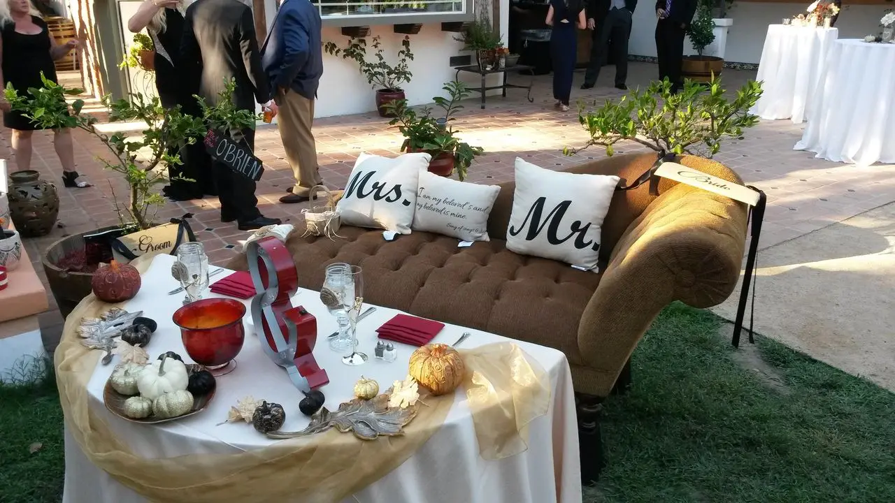 Wedding dj in los angeles A decorated outdoor wedding table set for two with "Mrs." and "Mr." pillows on a brown loveseat, surrounded by small pumpkins and autumn-themed decor. People are standing in the background.