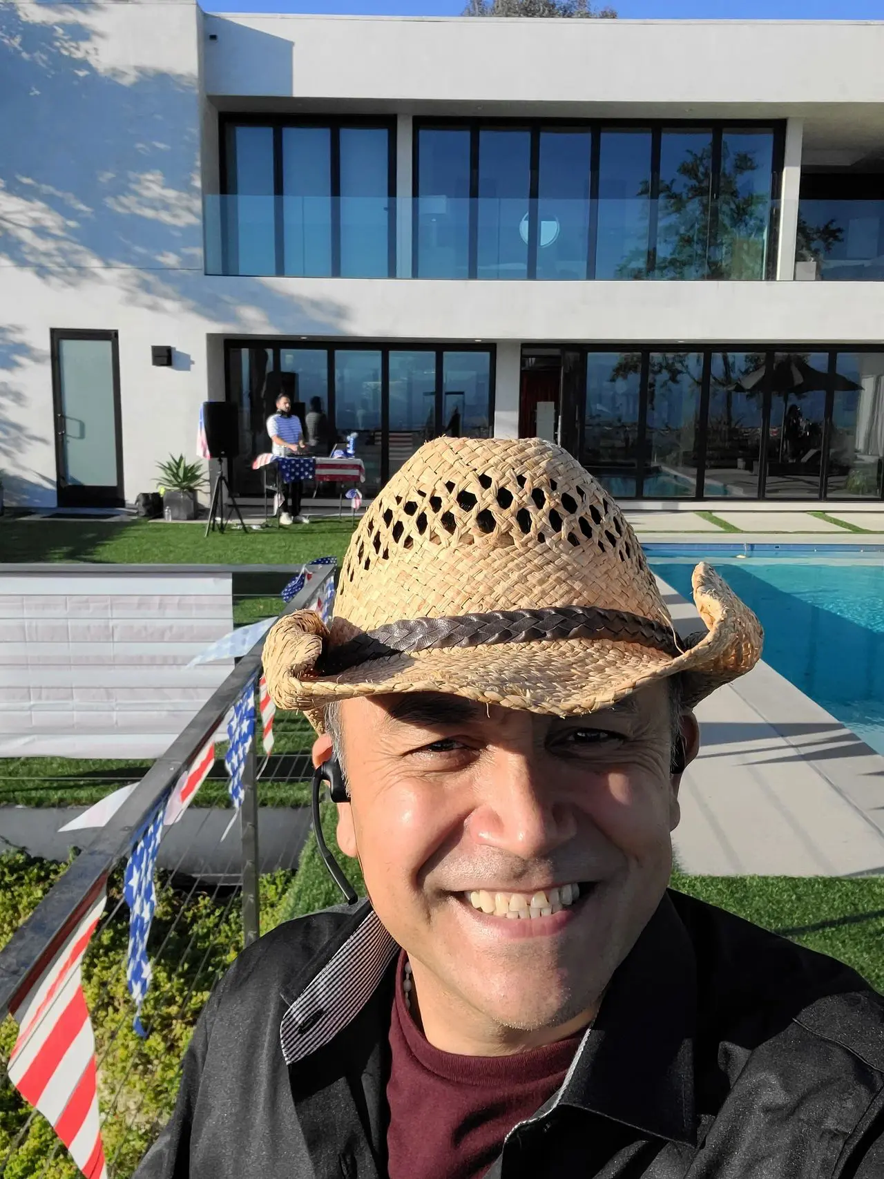Mr Dj Services - Photo Booth A man wearing a straw hat smiles for a selfie in front of a modern house with large windows, a pool, and outdoor decorations.