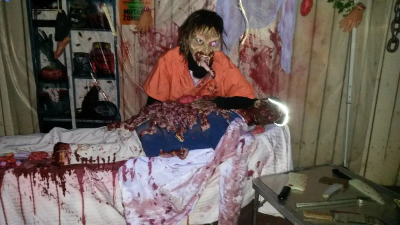 Halloween Dj Party A zombie prop in an orange shirt appears to eat fake intestines from a bloodied mannequin on a table, surrounded by Halloween decorations and props.