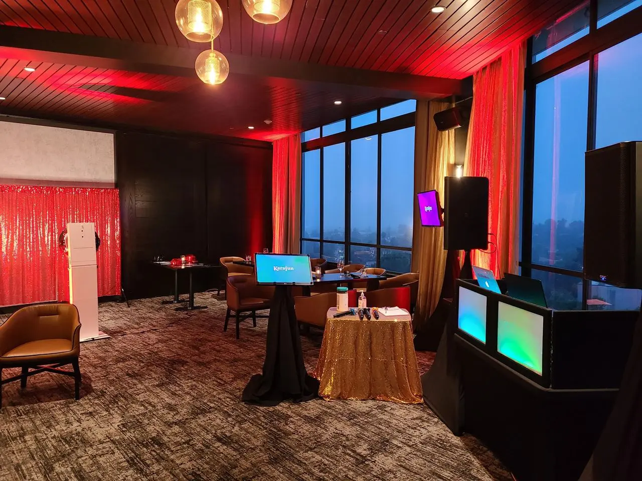Karaoke Dj Photo Booth Party A modern lounge with floor-to-ceiling windows, red ambient lighting, small tables, chairs, a DJ booth, and digital screens set up for an event.