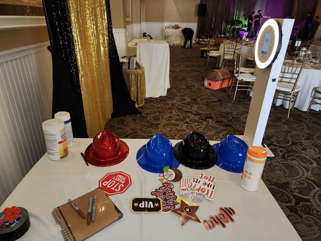 Photo Booth Fun A table with colorful plastic hats, photo booth props, wipes, and a guest book, next to a ring light photo booth at an event venue with empty chairs and tables.