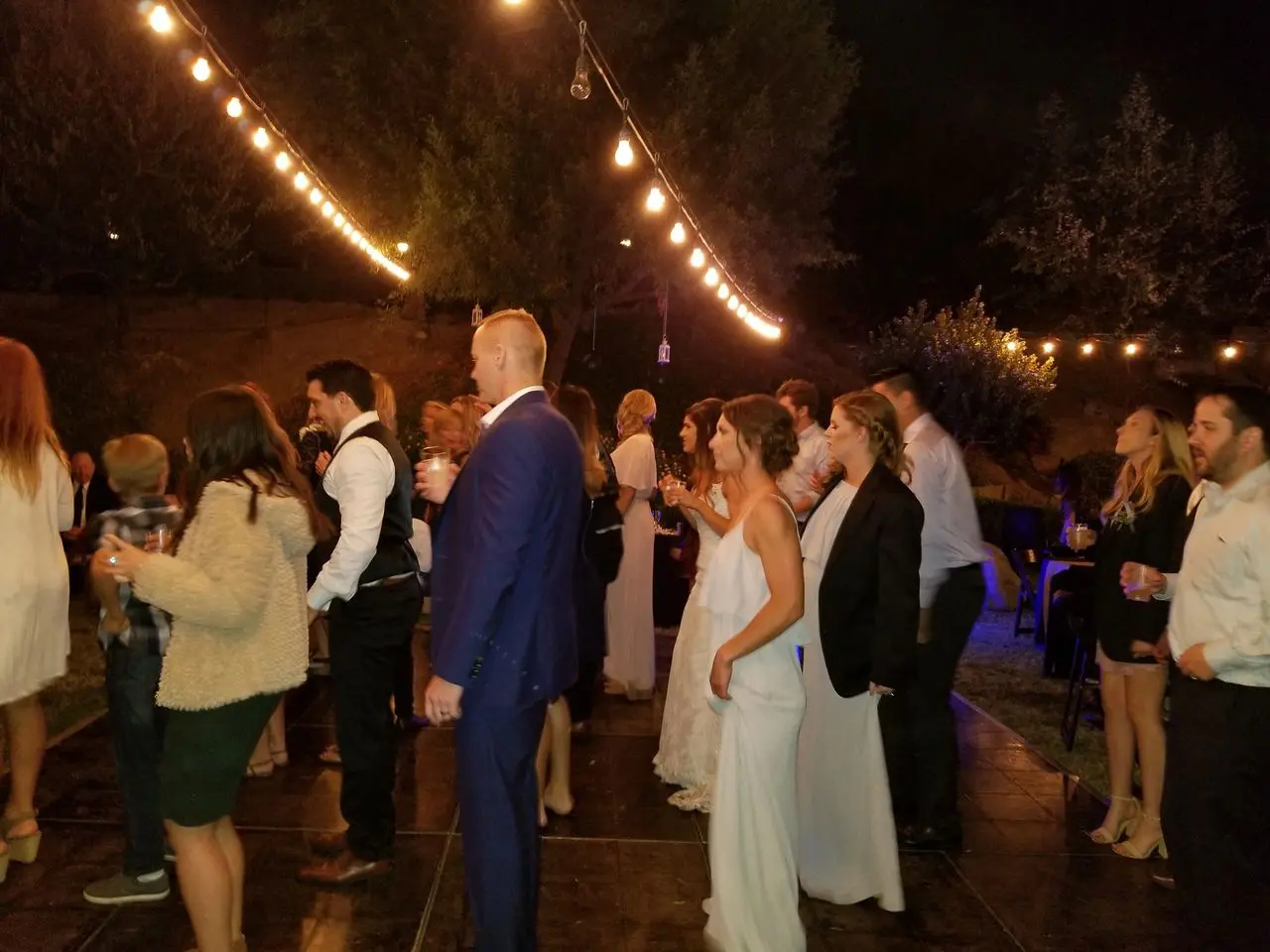 wedding dj in los angeles A group of people, some dressed formally, stand and dance under string lights at an outdoor nighttime event.