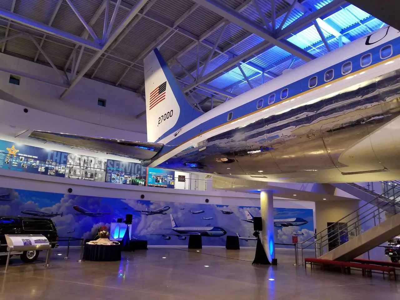Regan National Library A polished aircraft with tail number 21000 and a U.S. flag is displayed indoors at a museum, with exhibits and murals of planes and Secret Service memorabilia in the background.