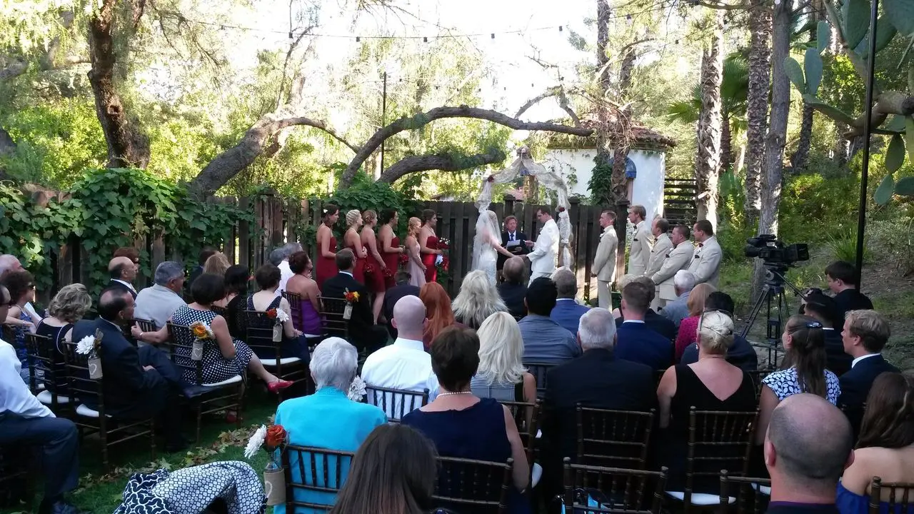 Mr Dj Services - wedding event dj planner Outdoor wedding ceremony with a bride and groom at the altar, guests seated on chairs, and bridesmaids in red dresses and groomsmen in light suits. Trees and greenery in the background.