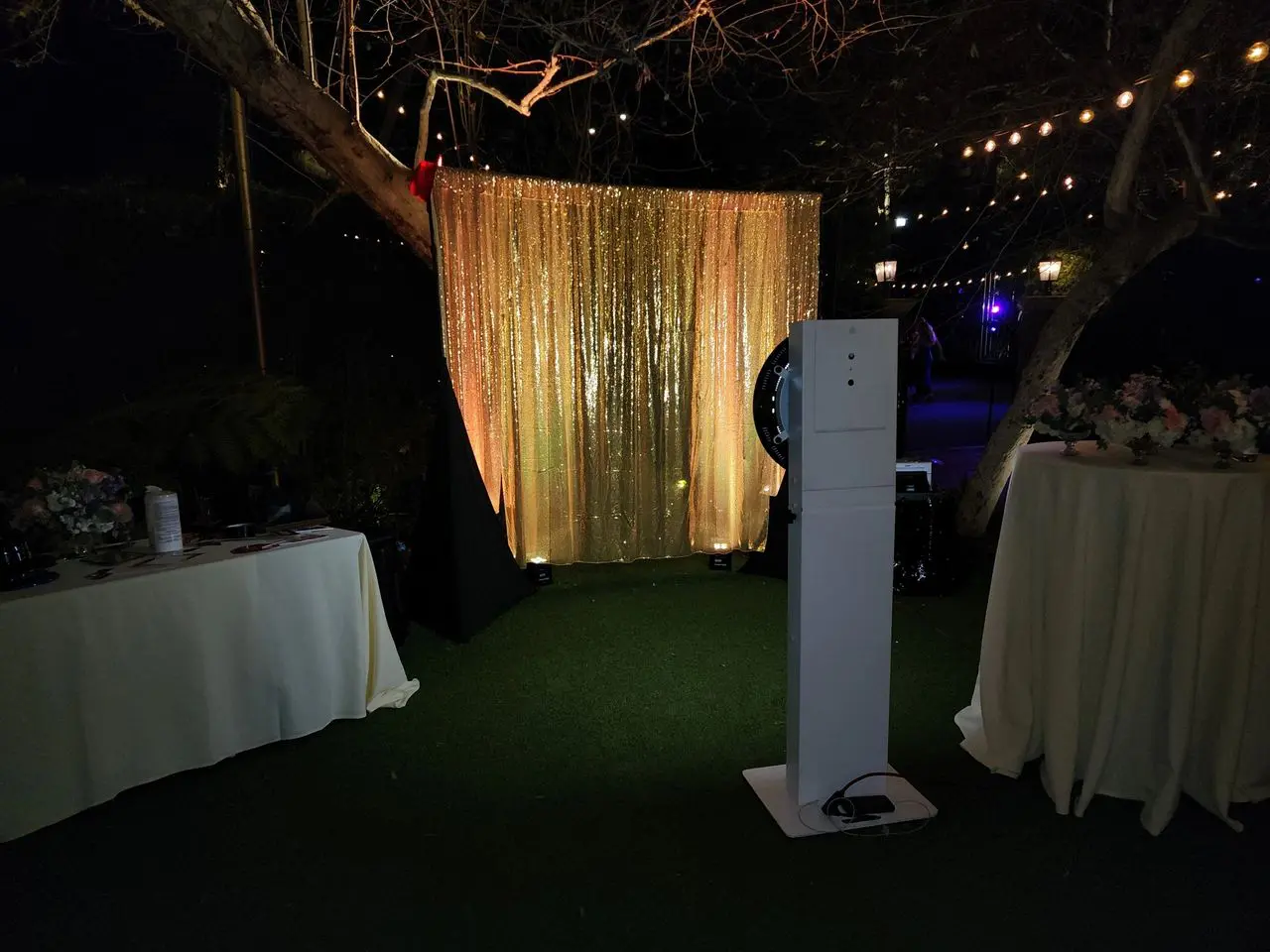 Mr Dj Services photo booth party An outdoor photo booth setup with a gold sequin backdrop, a camera stand in front, and two round tables covered with white cloths and floral arrangements. String lights hang above.