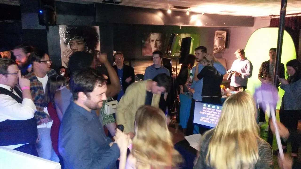 Mr Dj Services Wrap Karaoke Party A group of people gather and socialize in a dimly lit room with a green screen and karaoke machine, while a few hold microphones and sing.