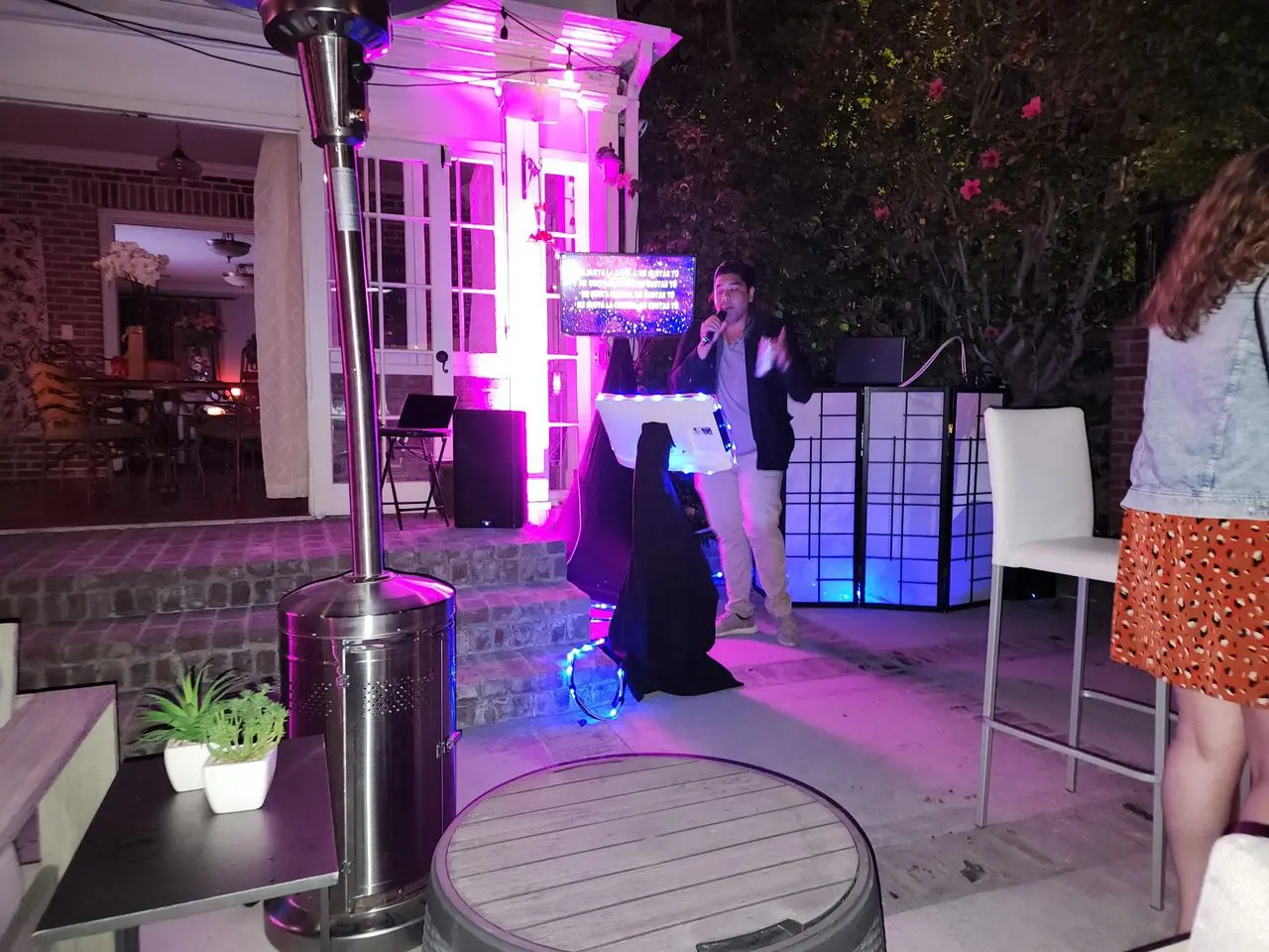 Mr Dj Services - Karaoke Dj Party A person sings into a microphone at an outdoor nighttime event with colorful lights, audio equipment, and several people in the background.