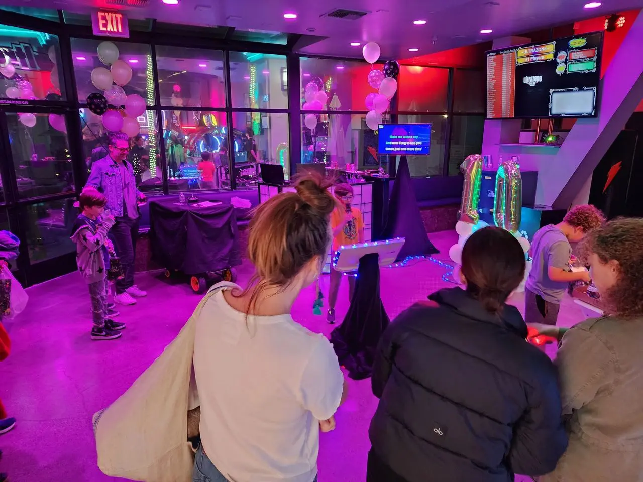Mr Dj Services People gathered in a neon-lit room decorated with balloons; a child sings karaoke while others watch or interact with games and activities.