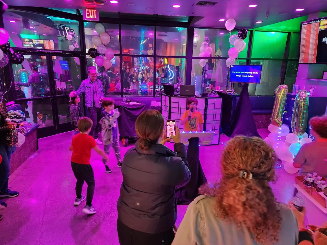 Mr Dj Services - Karaoke Dj Photo Booth Party Children dance at a birthday party in a room with purple lighting, balloons, and a DJ booth while adults watch and take photos.