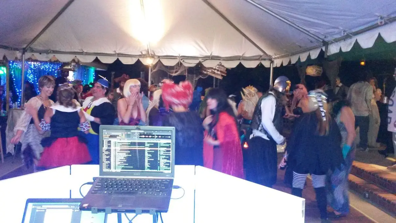 Halloween Dj Party A group of people in costumes are dancing under a tent at a party, viewed from behind a DJ booth with a laptop and equipment.