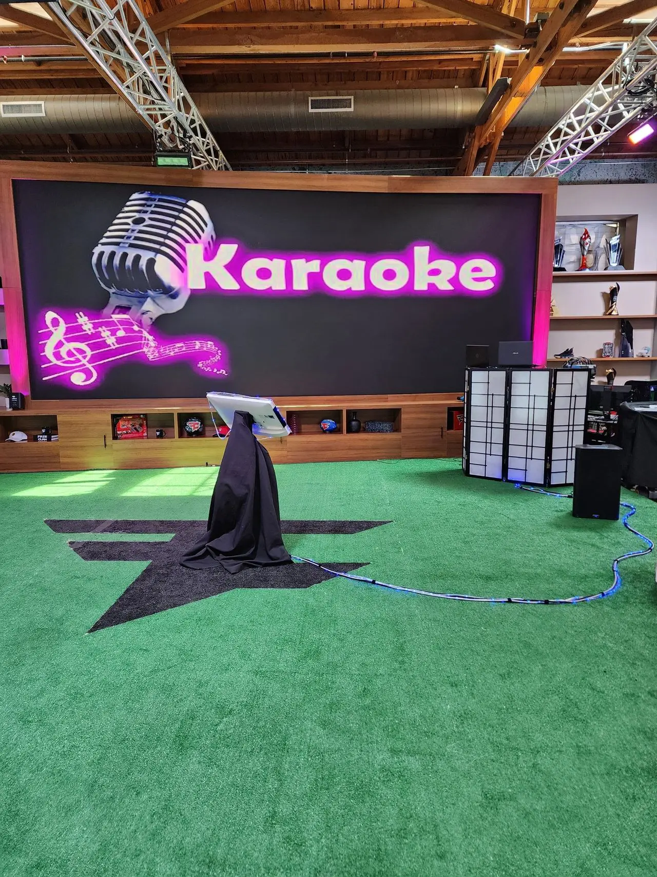 Corporate Karaoke Dj Party A karaoke setup with a microphone stand and screen on green turf in front of a large display that reads "Karaoke" with musical notes and a microphone graphic.