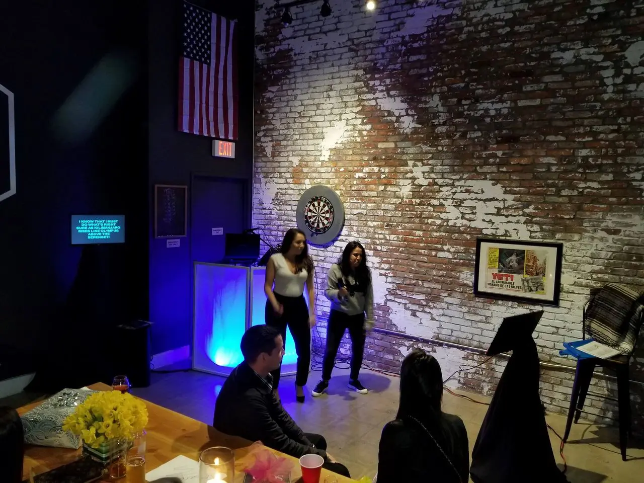 rs=w_1280 (7) Two women perform karaoke in a dimly lit room with a brick wall, dartboard, and American flag; several people are seated and watching.
