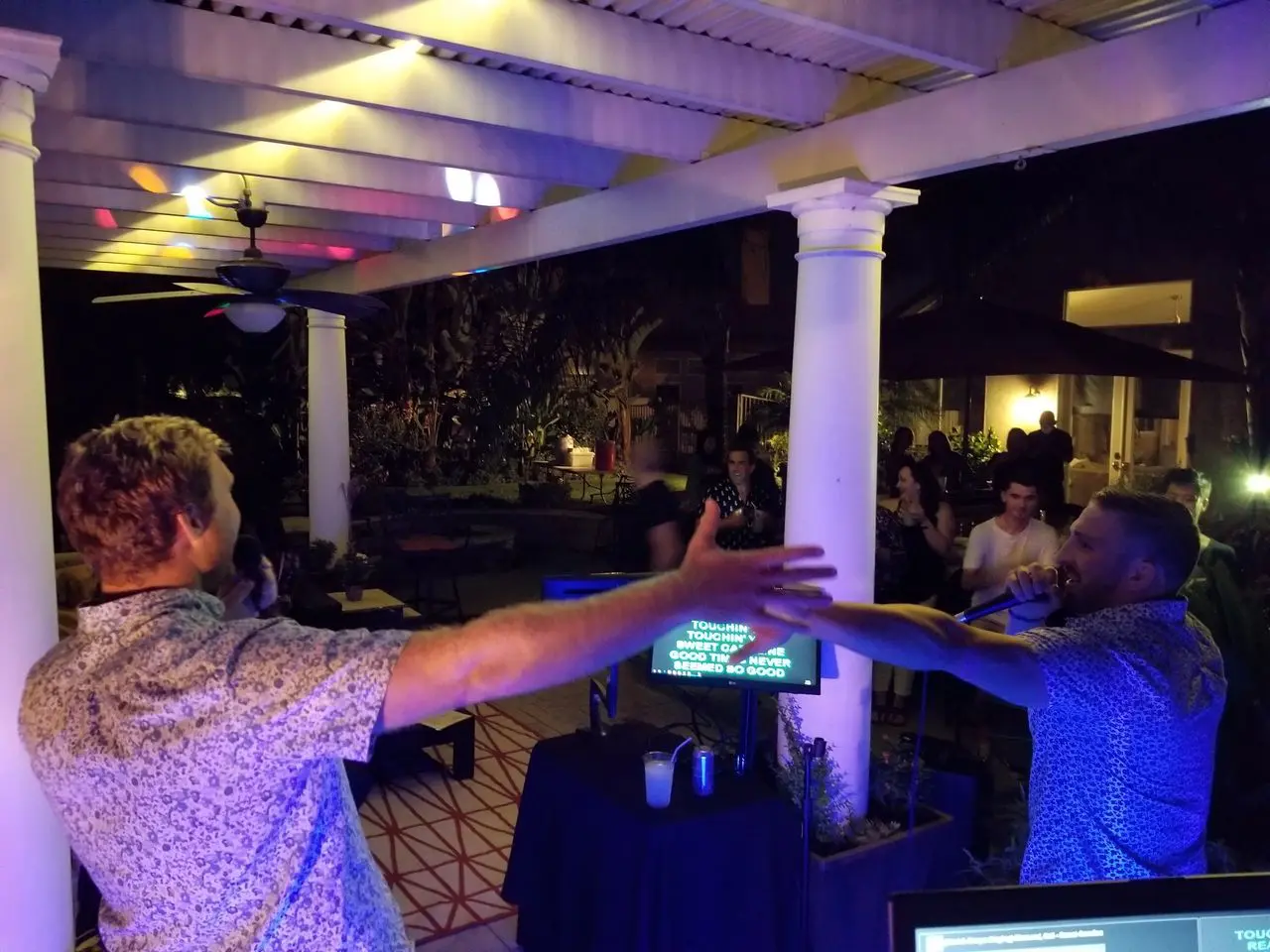 karaoke dj services spring Two men sing karaoke with microphones on an outdoor patio at night, facing each other with arms outstretched, while people watch in the background.