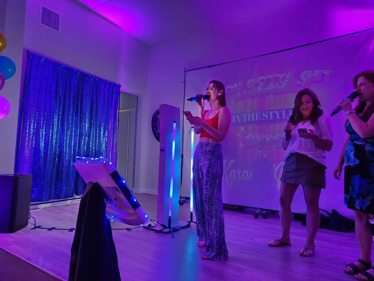 Karaoke Dj Mr Dj Services Three women sing karaoke on a small stage with purple lighting, holding microphones. A screen behind them displays song lyrics. Balloons and a sequin curtain are visible to the left.