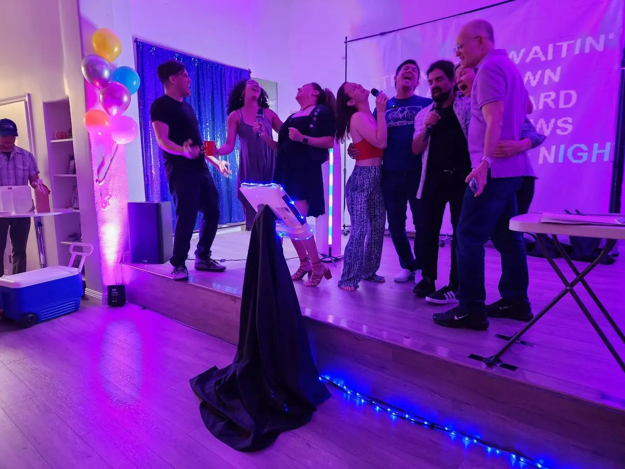 Happy bUrPdAy Birthday A group of people sing together on a small stage at a karaoke event, surrounded by colorful lights, balloons, and a projection screen in the background.