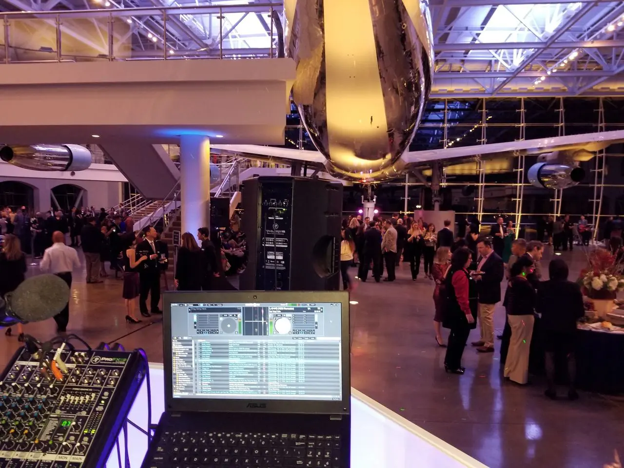 Dj for the President Regan A DJ setup with a laptop and mixer overlooks a crowd of people mingling at an indoor event space with a large airplane suspended overhead.