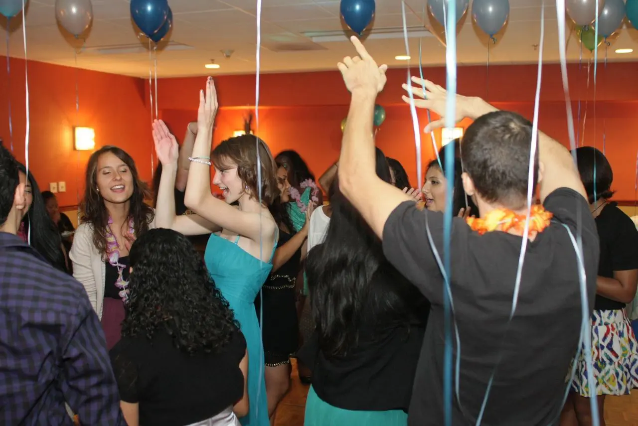 A group of people are dancing and celebrating in a decorated indoor room with balloons and orange walls.