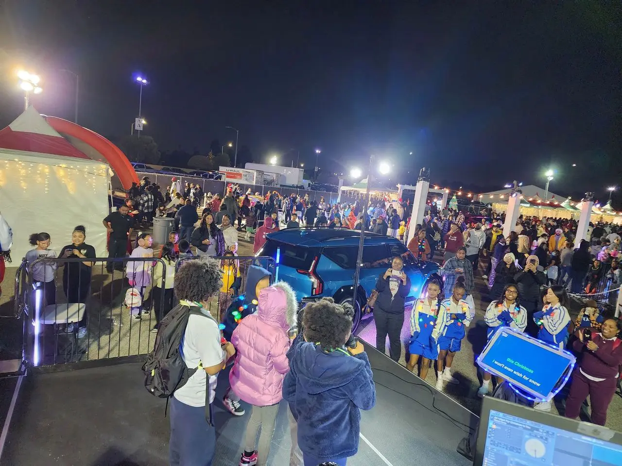 rs=w_1280 (8) Large crowd at a nighttime outdoor event with tents, a blue car, performers on stage, and various people walking and gathering under bright lights.