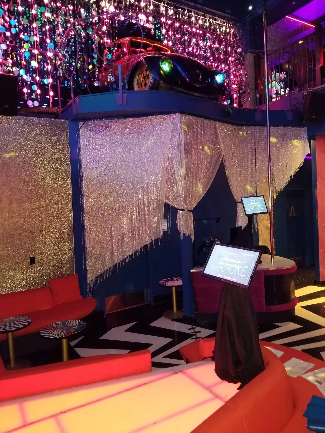 Mr Dj Services Electric Pussycat A brightly lit karaoke lounge with red seating, a stage, a karaoke machine, glittery decor, and a car displayed on a mezzanine level above.