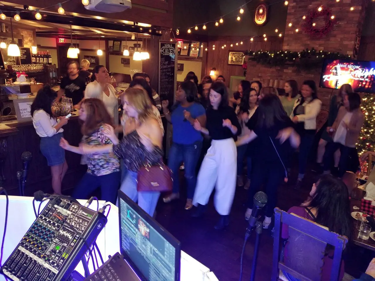 rs=w_1280 A group of people dance together in a warmly lit bar or restaurant, with music equipment visible in the foreground and festive decorations in the background.