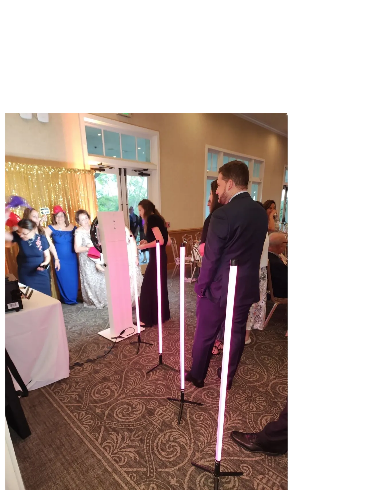 People dressed formally stand near a photo booth with pink lights at an indoor event. A gold sequin backdrop and props are visible in the background.