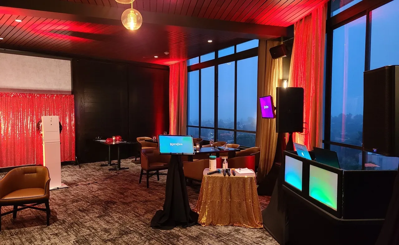 A modern event space with red lighting, tables, chairs, a sequin backdrop, two display screens, audio equipment, and large windows showing an evening city view.