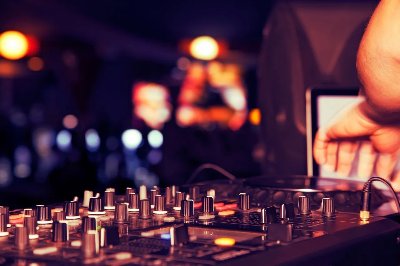 Close-up of a DJ mixing console with multiple knobs and sliders, a hand adjusting controls, and blurred colorful lights in the background.