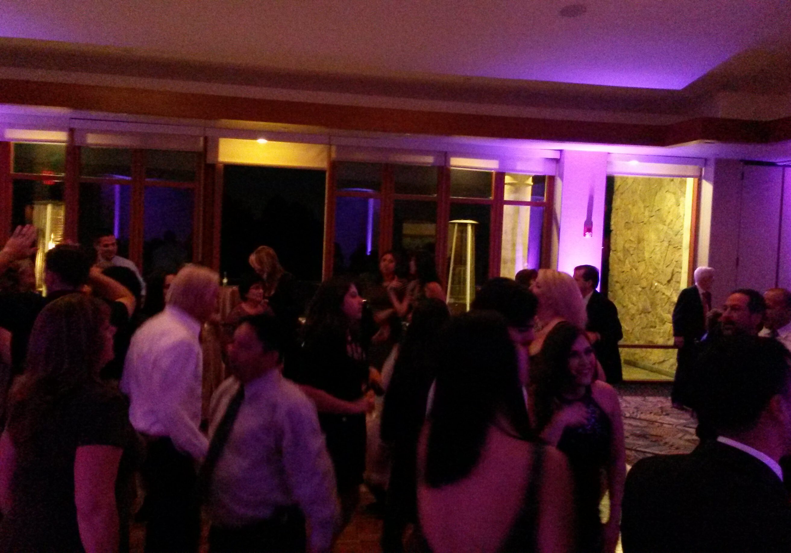 A group of people are dancing and socializing in a dimly lit room with purple lighting and large windows in the background.