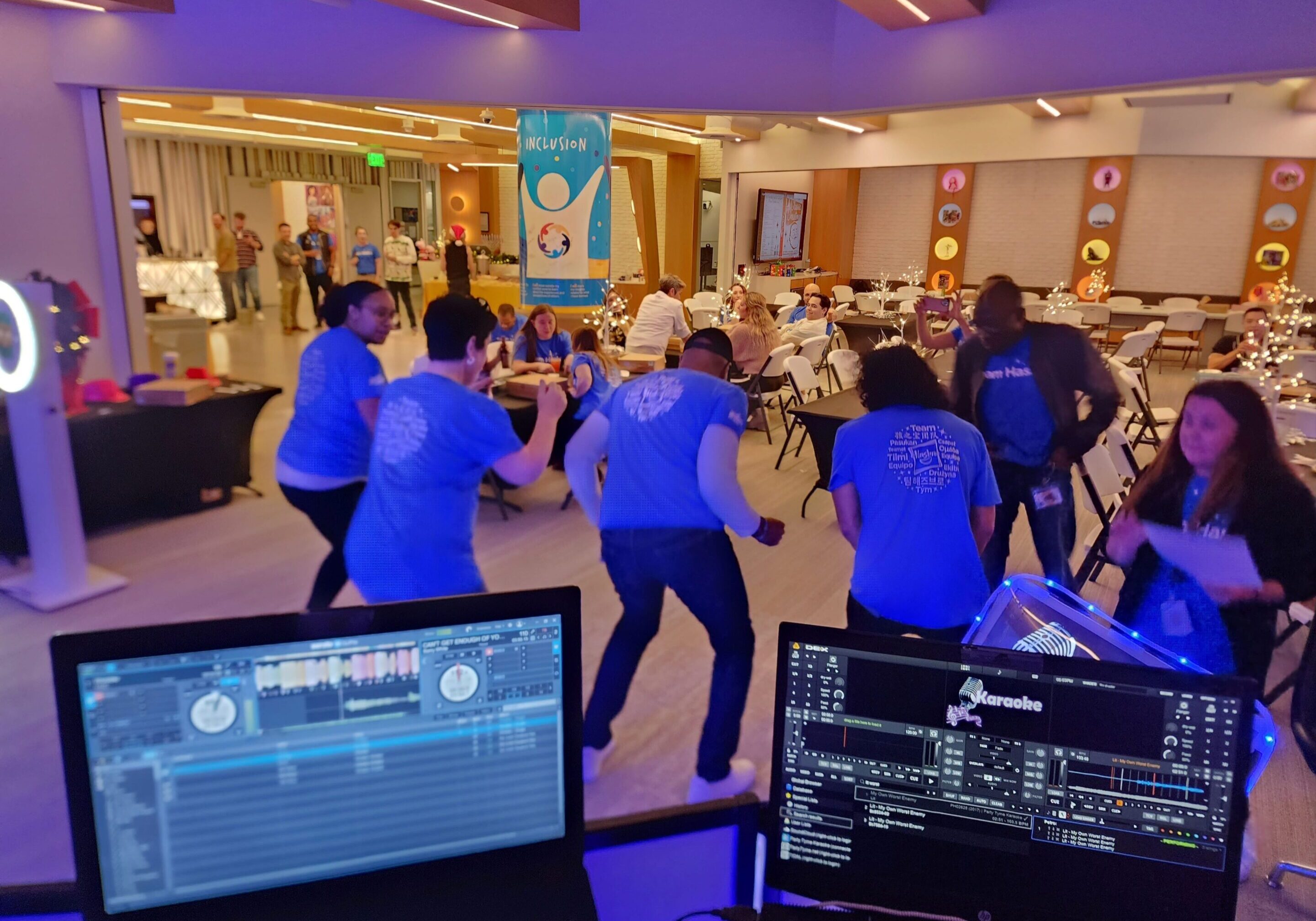 People dance in a brightly lit room during an indoor event, viewed from behind a DJ station music equipment and the song "wobble"