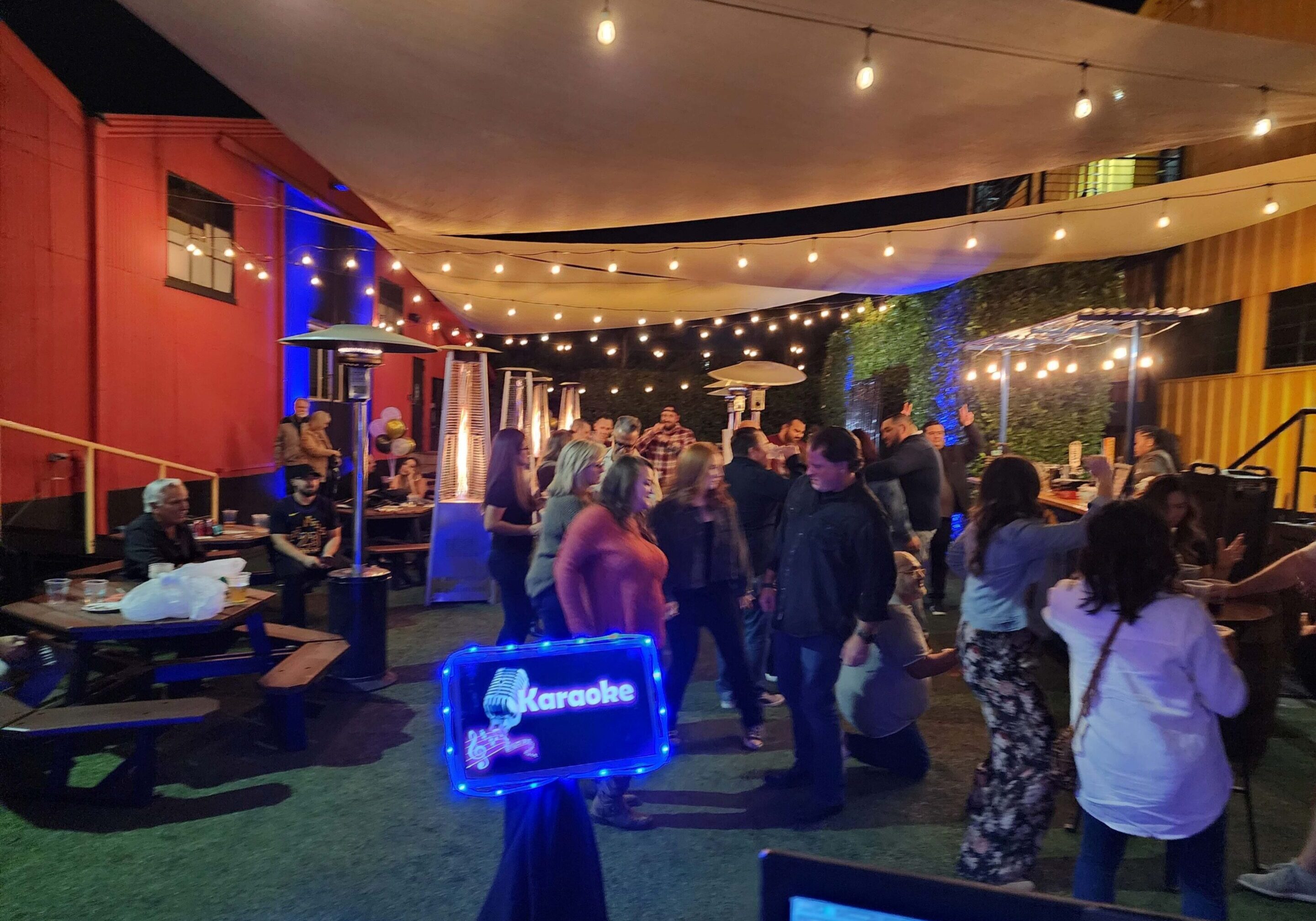 People gather outdoors under string lights and canopy for a nighttime karaoke event; some are seated at picnic tables, others stand and socialize.