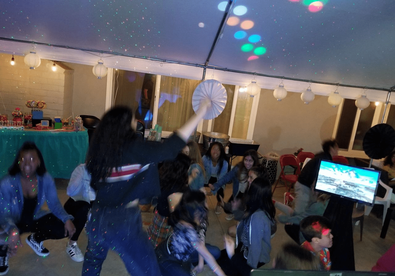 A group of people, mostly children, dance under a tent with string lights and colorful disco lights at a party. A decorated table and large paper fans are visible in the background.