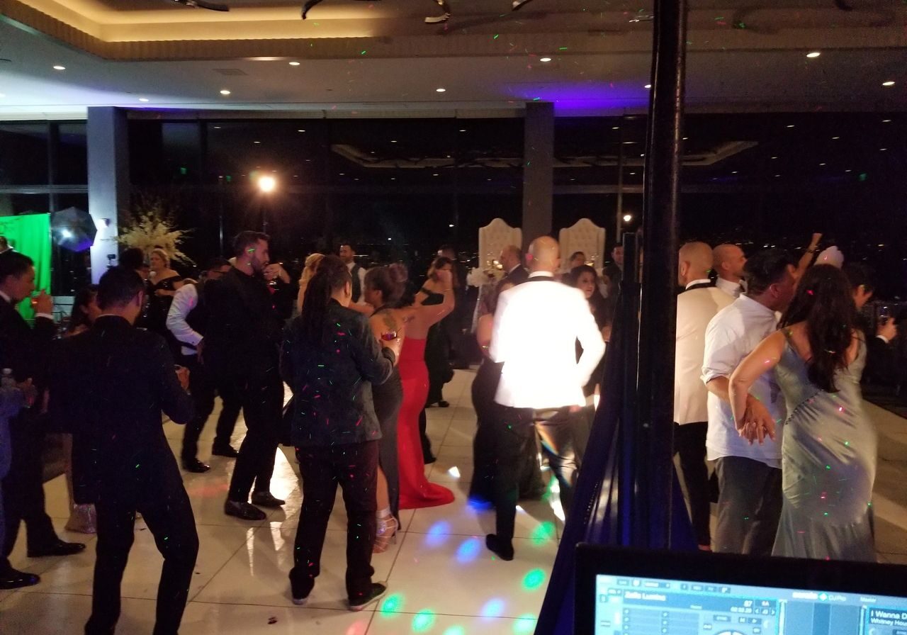 A group of people dressed in formal attire are dancing at an indoor event with dim lighting and colorful spotlights.