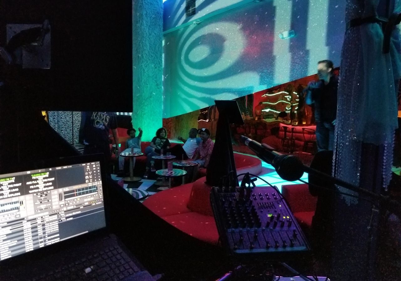 A DJ booth with a sound mixer and laptop overlooks a lounge where people are seated on red sofas while a person speaks or sings onstage under colorful lights.