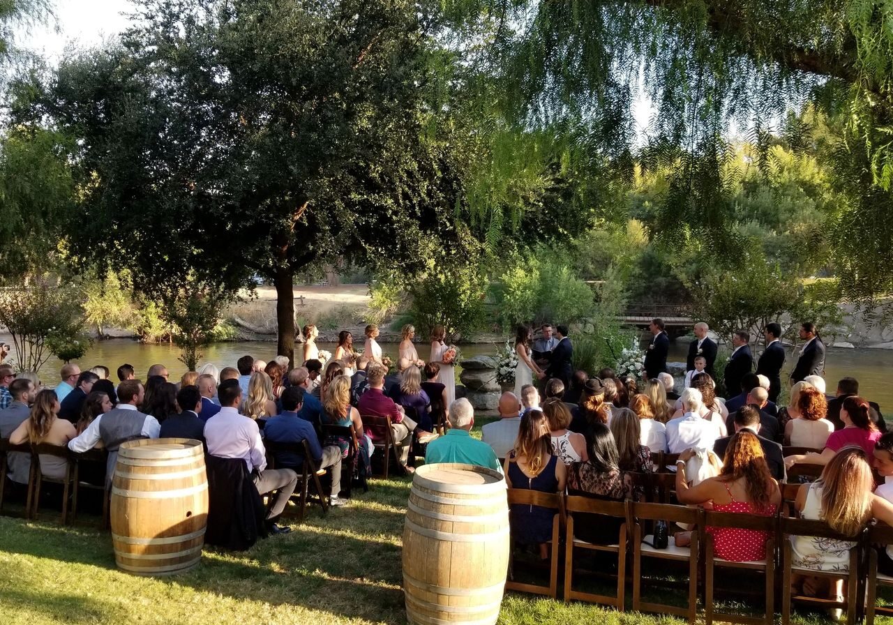 Outdoor wedding ceremony by a river with guests seated on wooden chairs, a wedding party at the front, and wine barrels in the foreground under trees.