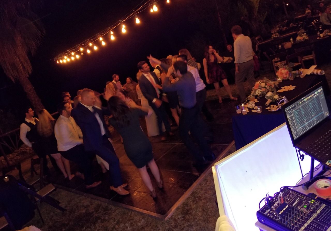 A group of people dance on an outdoor dance floor at night under string lights, with a DJ setup and sound equipment visible in the foreground.