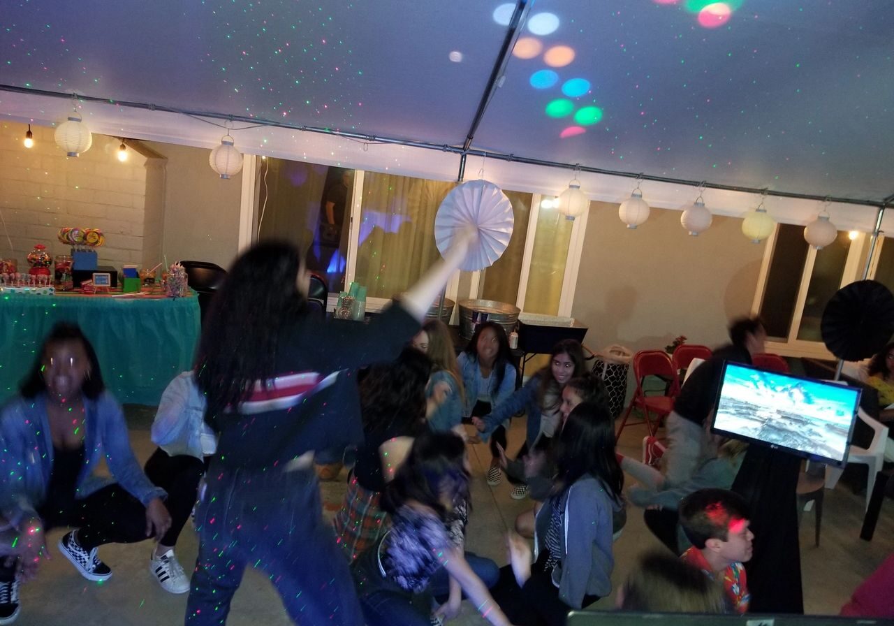 A group of people dance under colorful lights at an indoor party, with decorations and a table of snacks in the background.