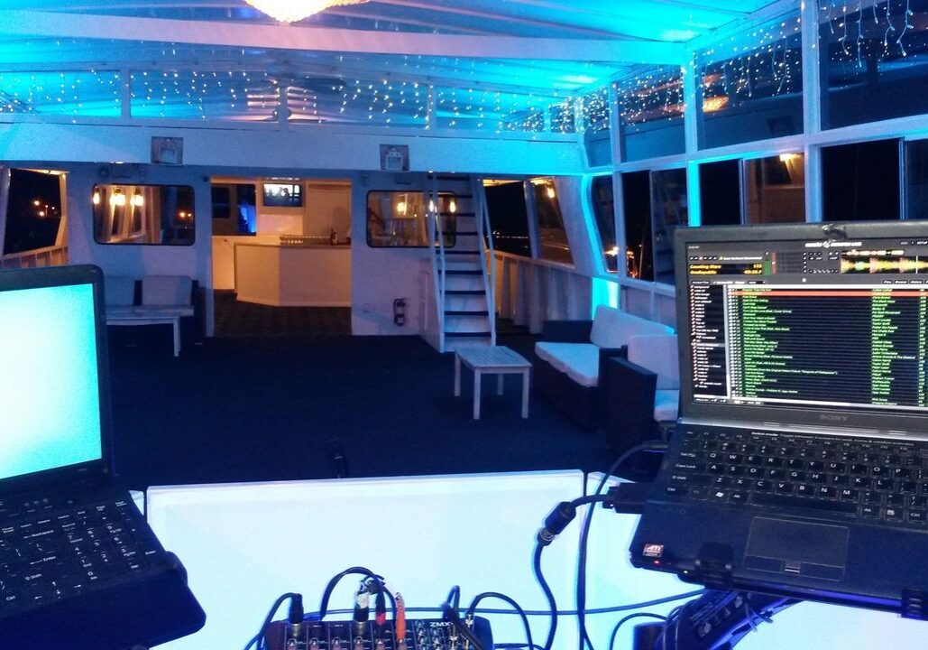 A DJ setup with two laptops and a mixer sits at the front of a brightly lit event space on a boat, featuring blue lighting, benches, and string lights on the ceiling.