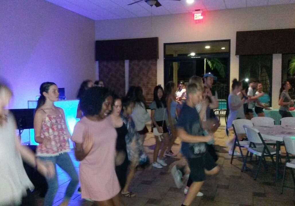 A group of people are dancing indoors in a well-lit room with tables and chairs set up around them.