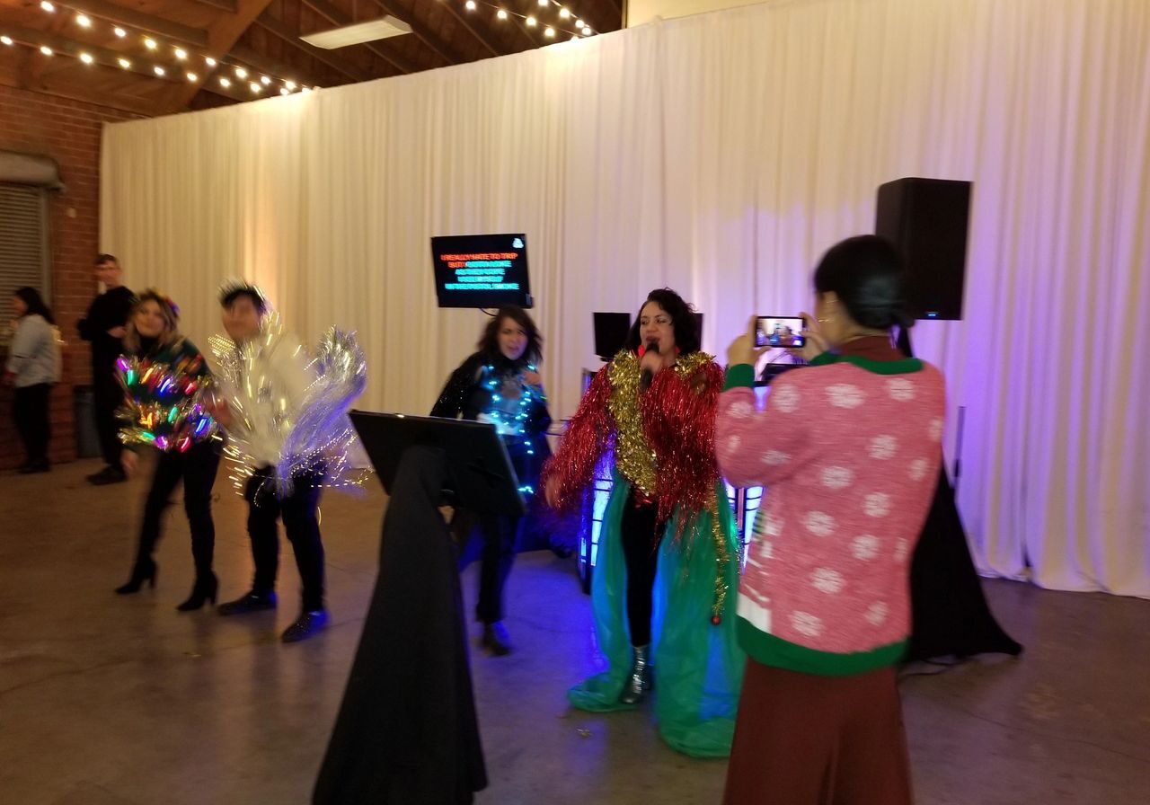 Four people perform karaoke in festive, shiny costumes while another person in a sweater films them with a phone in a room decorated with string lights and white curtains.