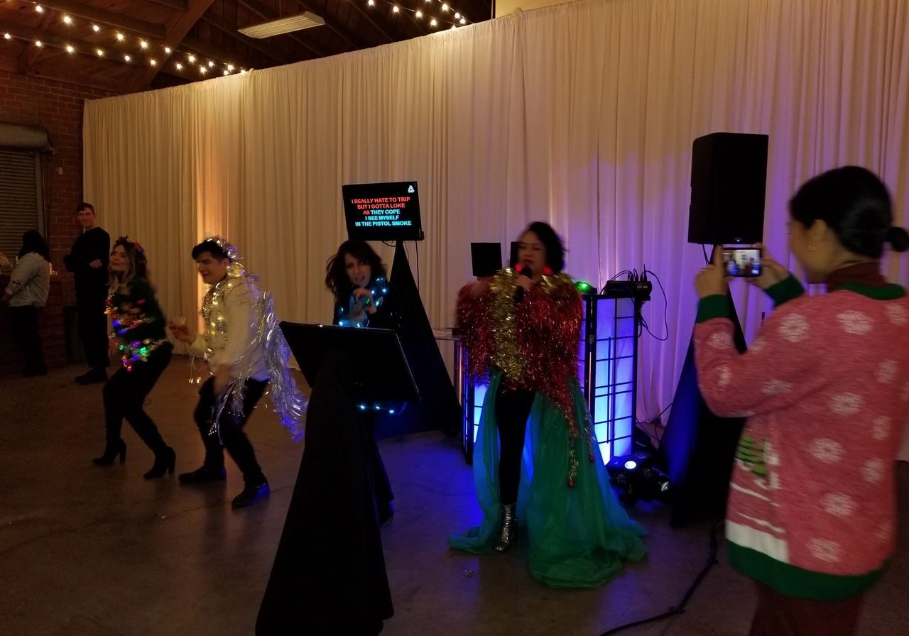 Four people in colorful, shiny costumes perform karaoke on a stage, while a woman in a festive sweater takes a photo with her phone. White drapes and string lights decorate the venue.