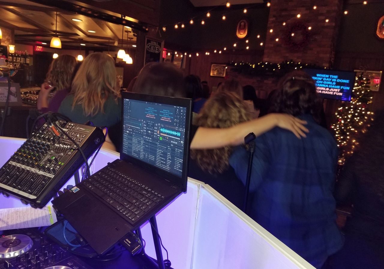 A group of people with arms around each other sing karaoke at a bar, viewed from behind a DJ booth with audio equipment and a laptop.