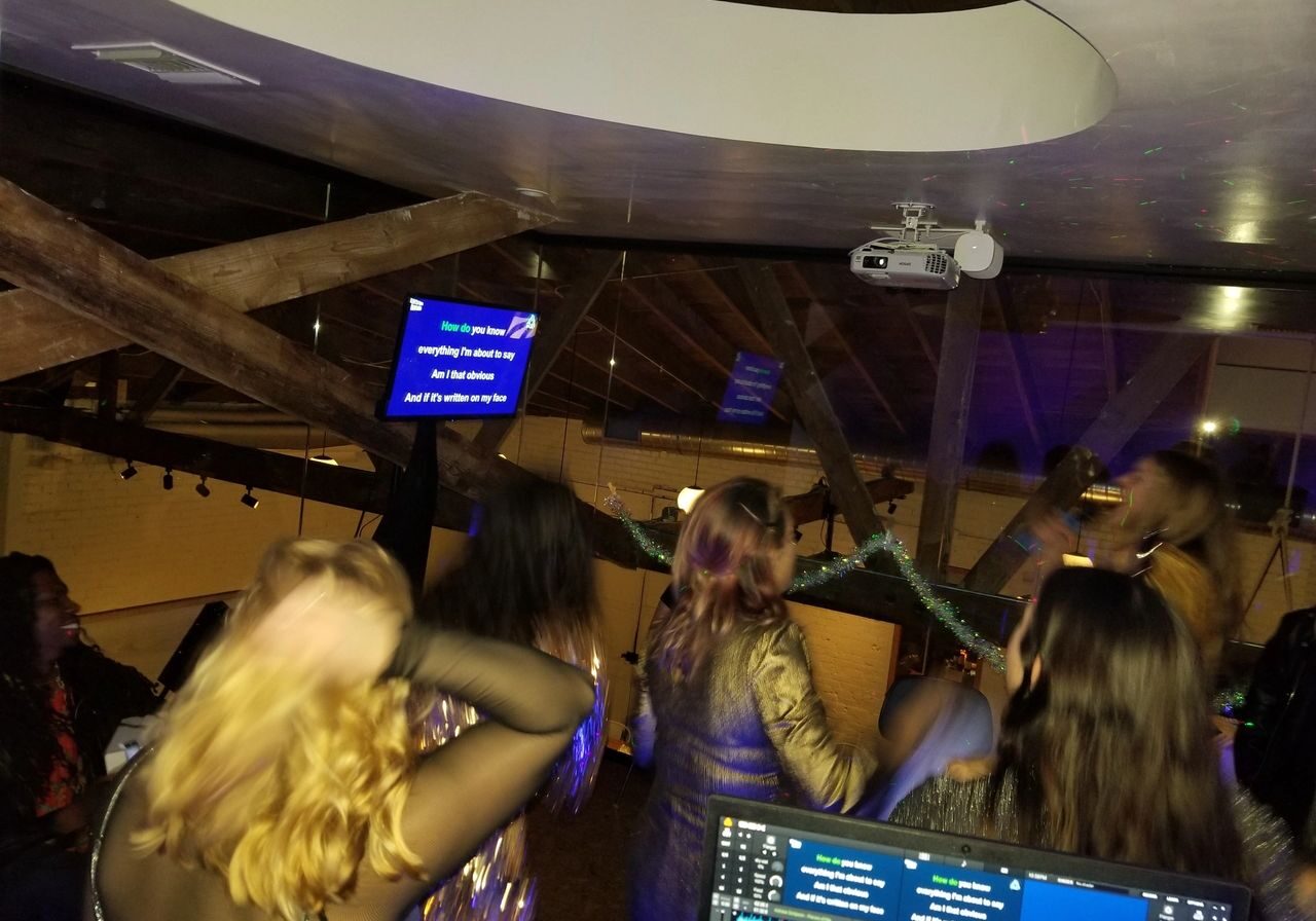 A group of people sing karaoke in a dimly lit room, facing a screen displaying song lyrics. A projector and DJ setup are visible in the background.