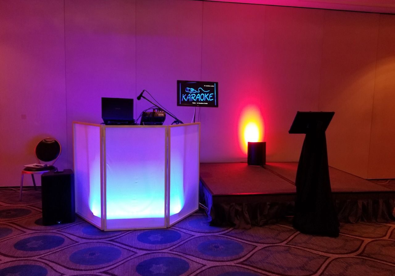 A karaoke setup with a lit DJ booth, microphone, laptop, monitor displaying “KARAOKE”, and a small stage with a music stand in a room with patterned carpet.
